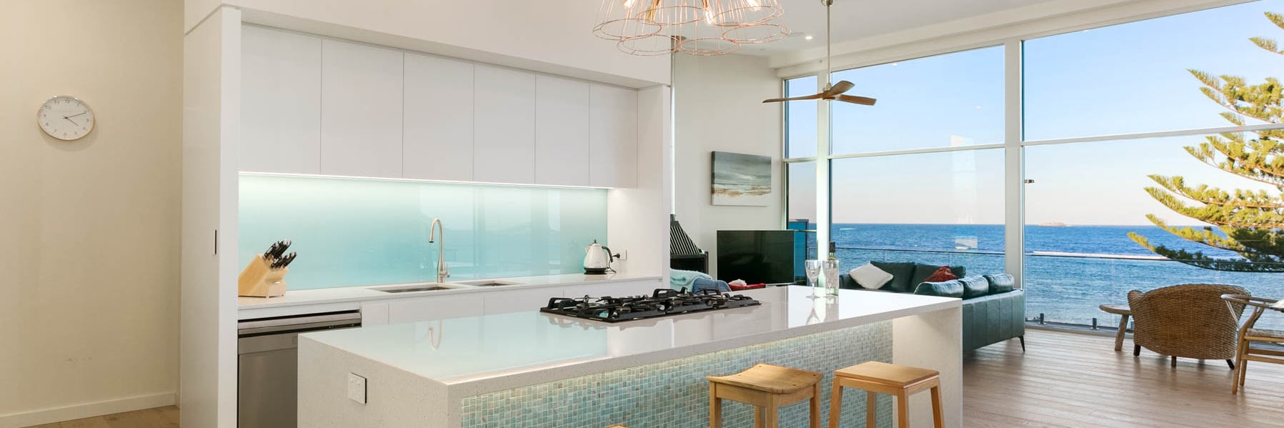 White cabinets with a motarised glass splashback. In front is a white island with integrated cooker top.