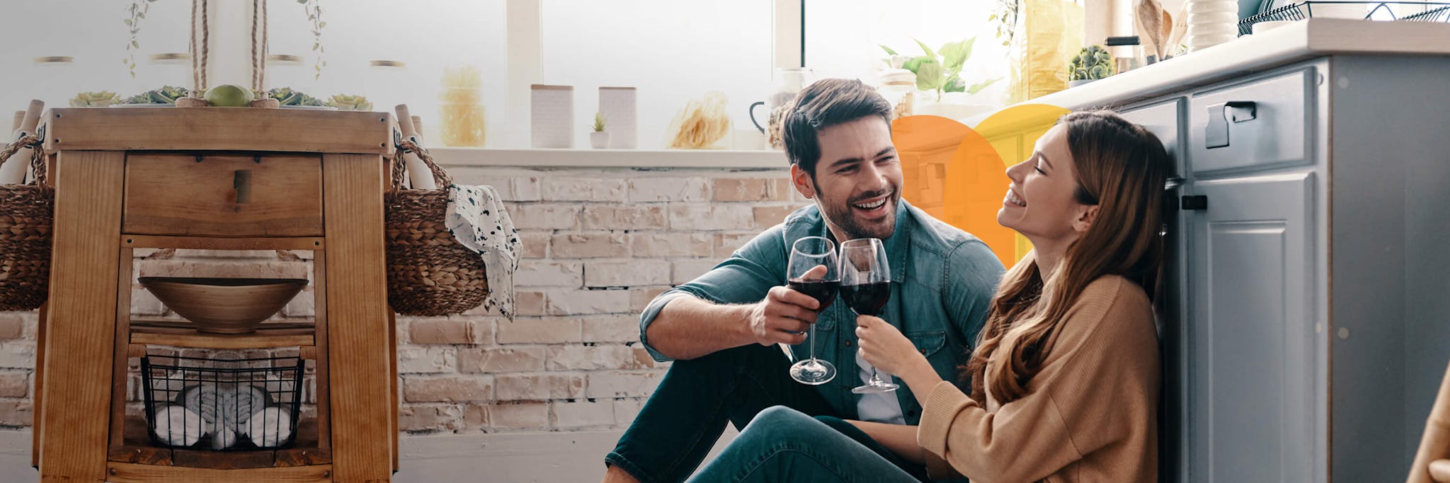 A couple sat on their kitchen floor smiling and drinking wine